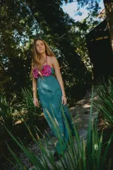 Vestido largo de tejido con lúrex color verde azulado, con escote recto y detalle de flores tipo rosette en color fucsia en el frente. Presenta un diseño con desniveles, tajo lateral y breteles finos.