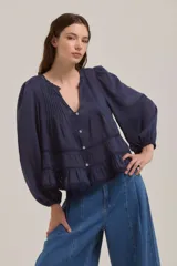 Camisa de color azul marino con cuello redondo y escote en V, mangas largas abullonadas con puños elásticos. Presenta un diseño con pliegues verticales en el pecho, detalles de encaje y volados en la parte inferior, con cierre frontal de botones.