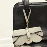 Estuche para lentes de diseño minimalista, confeccionado en cuero sintético texturizado color gris claro. Cuenta con cierre mediante broches a presión dorados y una correa superior con gancho metálico para colgar en bolsos o carteras.
