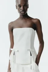 Top strapless de satén color blanco, con diseño entallado y detalle de dos lazos decorativos en el frente. Presenta aberturas en el bajo y cierre posterior con cremallera.