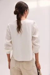 Blusa blanca de lino con escote en V y mangas largas con puños elásticos, combinando delantera estructurada de lino y espalda de algodón con spandex.