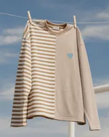 Buzo oversize de corte asimétrico con diseño combinado de rayas verticales y horizontales en tonos beige y blanco. Presenta un corazón bordado en color celeste en el frente y una etiqueta decorativa en la manga.