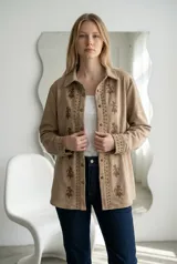 Sobrecamisa de gamuza color beige con diseño de bordados florales y detalles de tachas metálicas a lo largo de la botonadura frontal y en los puños. Presenta cuello clásico, cierre frontal con botones a presión y mangas largas.