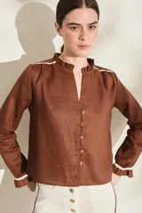 Blusa de lino color marrón con escote en V, cuello con volados y mangas largas con puños fruncidos que presentan detalles en color crudo, al igual que en los hombros. Cuenta con cierre frontal de botones.