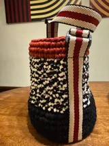 Bolso tejido a crochet con hilo de trapillo en colores negro, blanco y rojo. Tiene base de cuero vegano y correa de tela ajustable con detalles en rojo y blanco.