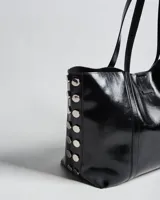 Cartera tote negra de símil cuero con acabado brillante, doble asa de hombro y detalles de tachas metálicas plateadas en los laterales.