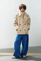Trench corto oversize color beige con cuello subido de efecto doble, cierre cruzado con botones oscuros y bolsillos de vivo delanteros.
