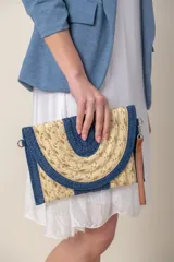 Sobre de mano o cartera tipo clutch, confeccionado en rafia tejida con detalles en lurex dorado y bordes en azul marino. Presenta un diseño frontal con forma de semicírculo en rafia natural y azul. Incluye una correa de muñeca desmontable de cuero sintético marrón.