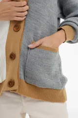 Cárdigan tejido de punto grueso, con diseño color block en gris, blanco y beige. Tiene cuello en V, cierre frontal con botones y un bolsillo de parche.