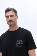 Remera de manga corta Brooksfield color negro, con cuello redondo y calce regular. Presenta un estampado frontal con la frase 'Into the nature' en letras cursivas azules y texto informativo debajo.