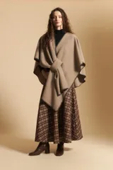 Poncho de tejido suave color beige con diseño envolvente y terminaciones con costuras decorativas en contraste color negro. Presenta un detalle de lazo frontal integrado.
