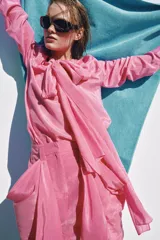 Blusa fluida de color rosado con cuello redondo ajustable mediante lazadas del mismo tejido, manga larga con puño y bajo asimétrico con aberturas laterales.