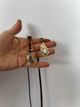 Collar artesanal con dije de base metálica irregular y piedra central roja. Presenta una estructura de tiento de gamuza marrón con detalles de tubos metálicos dorados.
