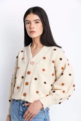 Cárdigan de lana color crudo con pompones marrones y naranjas, escote en V, cierre frontal con botones de madera, mangas largas abullonadas y corte holgado.