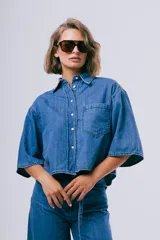 Camisa de denim azul de corte cropped, manga corta ancha, cuello camisero, cierre frontal de botones y un bolsillo aplicado en el pecho.