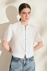 Blusa de lino color blanco, con cuello mao, cierre frontal con cremallera, mangas cortas abullonadas, pespuntes a contratono y un bolsillo aplicado en la parte inferior.