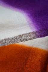 Bufanda tipo manta suave con diseño en bloques de color blanco, violeta y naranja, con flecos en los extremos.