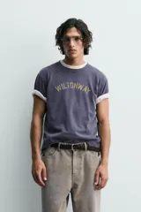Camiseta de manga corta, corte holgado, color gris oscuro o azul marino con efecto lavado y rotos. Estampado frontal con el texto "WILTONWAY" en letras doradas.