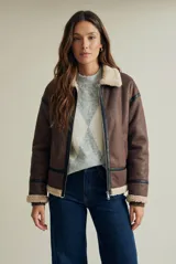 Campera tipo gamulán de corte oversize, color marrón con interior y cuello de corderito sintético en tono crema. Presenta detalles de ribetes en cuero sintético negro a lo largo de la prenda, cierre frontal metálico y puños tejidos.