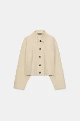 Trench corta de color beige, con cuello solapa, manga larga y cierre frontal con botones oscuros. Presenta bolsillos de vivo en el delantero y un diseño de corte recto.