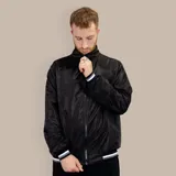 Campera bomber negra de nylon con cuello alto, cierre frontal con cremallera, bolsillos laterales con cierre y puños y cintura elásticos con rayas blancas, grises y azules.