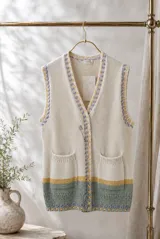 Chaleco largo de punto color crema con escote en V y cierre frontal. Presenta detalles bordados en tonos lila y amarillo en los bordes y sisas. Incluye dos bolsillos frontales y una franja inferior en tonos verde y amarillo.