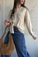 Camisa beige de manga larga con detalles bordados en el frente, cuello redondo y cierre frontal con botones. Las mangas presentan puños con volados.
