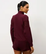 Campera deportiva tipo rompevientos color bordó, con cierre frontal, cuello alto y mangas largas. Presenta paneles laterales de tul para mayor ventilación y cordón ajustable en la cintura.