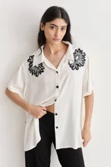 Camisa color crudo de corte amplio con bordados florales negros en la parte superior. Tiene cuello camisero, mangas cortas y cierre frontal con botones negros.