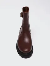 Bota de caña media en cuero color marrón oscuro, con suela track dentada y detalle de hebilla lateral en el tobillo.