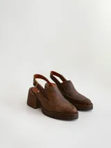 Zapato tipo zueco de cuero color chocolate con efecto desgastado, plataforma y taco cuadrado. Presenta una tira trasera ajustable con hebilla metálica.