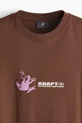 Remera de manga corta color marrón, con cuello redondo y corte holgado. Presenta un pequeño estampado en el pecho con la figura de una rana en color lila y la inscripción "ADAPTO OUTDOOR SUPPLIES".
