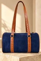 Cartera tipo baulito de cuero color marrón tostado, con doble asa larga de hombro, cierre superior y detalle de placa metálica con el logo de la marca en el centro.