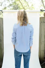 Camisa de popelina a rayas celestes y blancas, de estilo clásico, con cuello camisero, manga larga con puños ajustados, bolsillo frontal aplicado y botones de nácar.