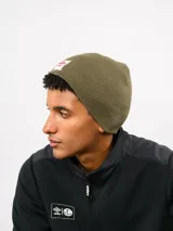 Gorro tipo beanie de tejido de punto acanalado en color verde oliva, con parche frontal rectangular blanco que presenta el logo de Umbro en rojo.