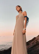 Vestido largo de diseño evasé en color beige, con cuello halter cruzado y espalda abierta. Presenta cierre de botones en el cuello.