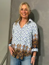 Blusa de manga larga con cuello camisero, corte recto y cierre frontal de botones. Presenta un diseño estampado geométrico en tonos celeste, blanco y marrón.