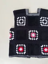 Chaleco sin mangas, confeccionado con cuadrados de cuero negro perforado combinados con cuadrados de crochet en hilo de algodón negro y blanco, con detalles centrales en rojo.