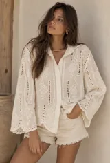 Camisa blanca de broderie con cuello camisero, cierre frontal con botones y mangas largas holgadas.