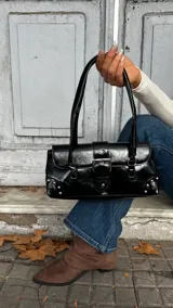 Cartera de hombro negra con acabado brillante, diseño rectangular con solapa frontal, cierre de hebilla metálica y detalles de tachas plateadas en los extremos inferiores.