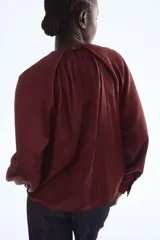 Blusa de satén color borgoña con mangas globo largas y puños anchos. Presenta un detalle de drapeado o nudo en el centro del escote redondo.