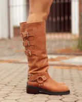 Bota de caña alta confeccionada en cuero color marrón tostado. Presenta un diseño con tres hebillas decorativas en la parte superior de la caña y una correa con hebilla y argolla metálica en el tobillo. Cuenta con suela plana y taco bajo de madera oscura.