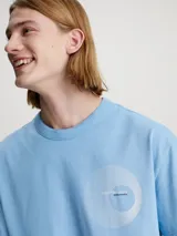 Remera de algodón en tono celeste claro, corte relajado y manga corta, con cuello redondo. Presenta un pequeño logo gráfico circular que simula un vinilo o disco en el lado izquierdo del pecho.