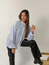 Camisa de rayas verticales celestes y blancas, con cuello alto y cierre frontal de botones. Presenta detalles de puntilla blanca en el cuello, a lo largo de la botonera y en los puños. Tiene mangas largas con terminación acampanada y diseño holgado.