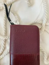 Billetera grande de cuero marca Rupia, con diseño bicolor en tono bordó. Presenta una textura granulada en la parte superior y una textura tipo reptil en la parte inferior. Incluye cierre metálico con tirador de cuero negro y un dije metálico circular plateado con el logo de la marca.