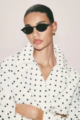 Cazadora estilo trench corta confeccionada en lino, con diseño de lunares negros sobre fondo blanco. Cuenta con cuello solapa, manga larga con trabillas en los puños, cierre frontal cruzado con botones negros y cinturón ajustable del mismo tejido.
