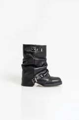 Botas de cuero de caña media con diseño slouchy, caracterizadas por su acabado arrugado y apliques de hebillas metálicas en la parte superior y en el tobillo. Cuentan con una suela plana y punta redondeada.