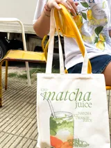 Bolso tote color beige claro con asas de tela, presentando un estampado gráfico de un vaso de jugo de matcha con capas de colores verde, blanco y naranja, y texto que dice "matcha juice".