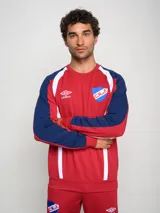 Buzo deportivo de manga larga, color principal rojo, con mangas azul marino y detalles en blanco que bajan desde el hombro hasta el puño. Presenta cuello redondo, logo Umbro y escudo del Club Nacional de Football en el pecho.
