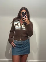 Campera de punto color marrón con cierre frontal, cuello alto y diseño estampado de caballos en la parte superior con franjas decorativas en blanco y celeste.